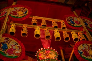 A vibrant display featuring handmade decorative items, including intricately painted circular fans with vivid, traditional patterns. Yellow cylindrical ornaments with hand-drawn figures hang from a wooden frame. The scene is filled with rich reds and golds, creating a festive and cultural atmosphere.