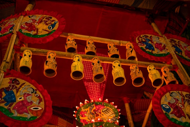 A vibrant display featuring handmade decorative items, including intricately painted circular fans with vivid, traditional patterns. Yellow cylindrical ornaments with hand-drawn figures hang from a wooden frame. The scene is filled with rich reds and golds, creating a festive and cultural atmosphere.
