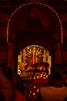 A Hindu deity is enshrined in a temple adorned with multiple intricate designs and colorful decorations. The main focus is on the central idol, which has multiple arms holding various objects. The shrine is illuminated with numerous small lights, creating a warm and spiritual ambiance. People are gathered in front of the shrine, likely engaged in worship or prayer.