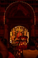 A Hindu deity is enshrined in a temple adorned with multiple intricate designs and colorful decorations. The main focus is on the central idol, which has multiple arms holding various objects. The shrine is illuminated with numerous small lights, creating a warm and spiritual ambiance. People are gathered in front of the shrine, likely engaged in worship or prayer.