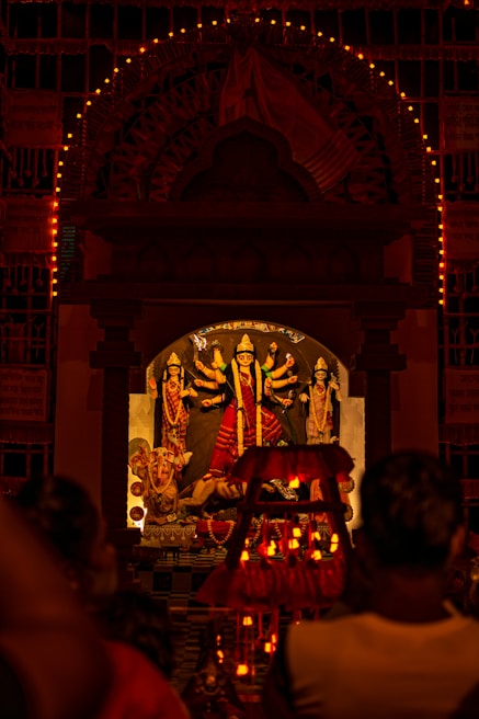 A Hindu deity is enshrined in a temple adorned with multiple intricate designs and colorful decorations. The main focus is on the central idol, which has multiple arms holding various objects. The shrine is illuminated with numerous small lights, creating a warm and spiritual ambiance. People are gathered in front of the shrine, likely engaged in worship or prayer.