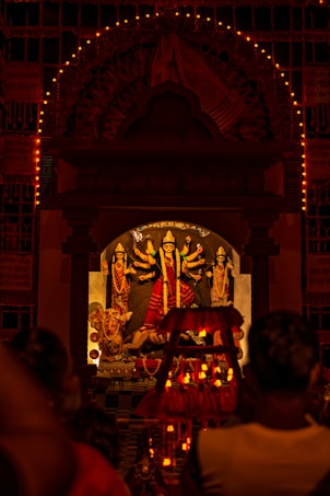 A Hindu deity is enshrined in a temple adorned with multiple intricate designs and colorful decorations. The main focus is on the central idol, which has multiple arms holding various objects. The shrine is illuminated with numerous small lights, creating a warm and spiritual ambiance. People are gathered in front of the shrine, likely engaged in worship or prayer.