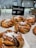 Freshly baked cinnamon buns are displayed on a tray with a backdrop of a kitchen setting. They are topped with white sugar crystals, and a small chalkboard sign indicates their name and price. The interior decor features white subway tiles and shelves with plates and cups.