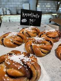 Freshly baked cinnamon buns are displayed on a tray with a backdrop of a kitchen setting. They are topped with white sugar crystals, and a small chalkboard sign indicates their name and price. The interior decor features white subway tiles and shelves with plates and cups.