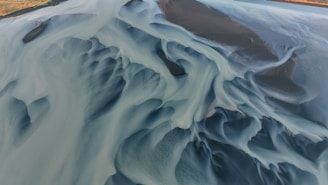 An aerial view of a river delta showcasing intricate, abstract patterns created by water channels and sediment. The image features a mix of flowing lines and smooth, curving shapes, with areas of varying depth and texture. The colors range from light blue and grey to darker, almost black tones, revealing the dynamic interplay between water, sediment, and the riverbed.