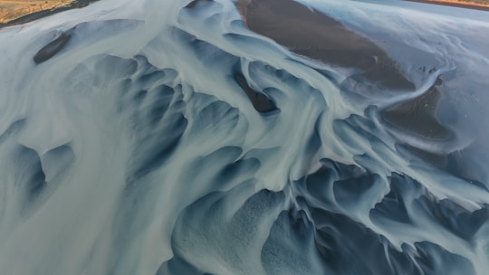 An aerial view of a river delta showcasing intricate, abstract patterns created by water channels and sediment. The image features a mix of flowing lines and smooth, curving shapes, with areas of varying depth and texture. The colors range from light blue and grey to darker, almost black tones, revealing the dynamic interplay between water, sediment, and the riverbed.