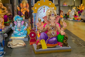 The image displays several colorful statues of the Hindu deity Ganesha, each adorned with intricate jewelry and vibrant attire. The central figure of Ganesha is sitting on a platform, holding various items in his hands, and is decorated with a crown and other traditional ornaments. Surrounding Ganesha are other similar statues in different poses and colors. The scene is set indoors, likely in a shop or workshop, with additional figures in the background.