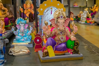 The image displays several colorful statues of the Hindu deity Ganesha, each adorned with intricate jewelry and vibrant attire. The central figure of Ganesha is sitting on a platform, holding various items in his hands, and is decorated with a crown and other traditional ornaments. Surrounding Ganesha are other similar statues in different poses and colors. The scene is set indoors, likely in a shop or workshop, with additional figures in the background.
