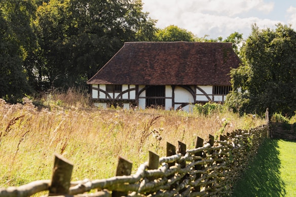 A picturesque half-timbered building with a dark pitched roof surrounded by lush greenery and tall grasses. The foreground features a rustic wooden fence leading towards the house. The scene is bathed in warm sunlight, casting gentle shadows and highlighting the textures of the materials.