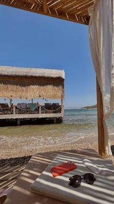 A serene beach setting with a thatched-roof cabana overlooking the sea. There is a sunbed with a phone in a red case, sunglasses, and sunscreen placed on it. The cabana provides shade while a gentle breeze moves the curtains. In the background, people can be seen near the shore, relaxing and enjoying the seaside view.