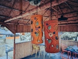 Two intricately designed hanging lamps with colorful floral patterns are the focal point in an open bamboo structure. The surrounding area features bamboo walls and furniture, including a table with assorted items and chairs.