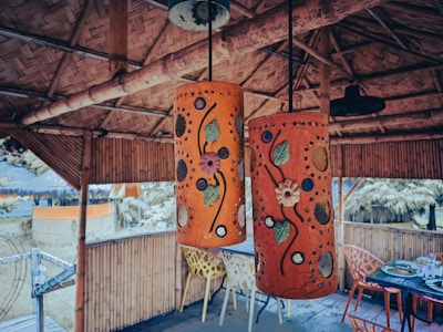 Two intricately designed hanging lamps with colorful floral patterns are the focal point in an open bamboo structure. The surrounding area features bamboo walls and furniture, including a table with assorted items and chairs.