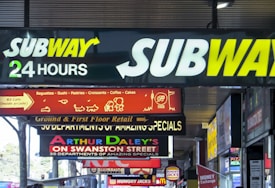 A busy urban street scene with numerous illuminated signs. Prominent among them is the Subway logo, indicating 24-hour service. Other signs advertise various dining options like baguettes, sushi, pastries, croissants, coffee, and cakes, as well as retail and special offers on Swanston Street. Bright lights and diverse fonts create a bustling city atmosphere.