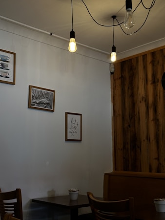 A cozy caf&eacute; corner with wooden accents and hanging lights. Framed pictures and a quote about coffee adorn the wall. A small table with chairs occupies the corner, and a potted plant adds a touch of greenery.
