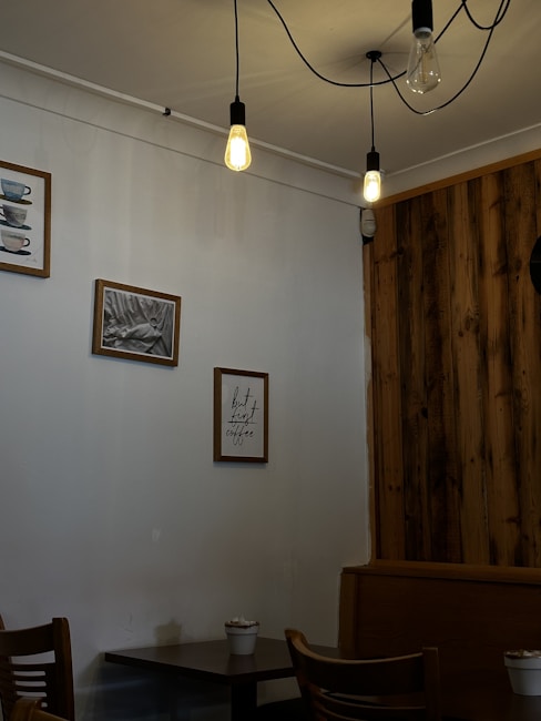 A cozy caf&eacute; corner with wooden accents and hanging lights. Framed pictures and a quote about coffee adorn the wall. A small table with chairs occupies the corner, and a potted plant adds a touch of greenery.