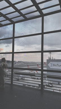 A large window grid frames an airport runway with multiple airplanes bearing Turkish Airlines livery. The scene includes a cloudy sky and airport infrastructure visible in the background. Two people stand near the window, gazing outside, with a sign for BtcTurk in the distance.
