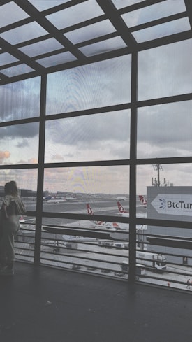 A large window grid frames an airport runway with multiple airplanes bearing Turkish Airlines livery. The scene includes a cloudy sky and airport infrastructure visible in the background. Two people stand near the window, gazing outside, with a sign for BtcTurk in the distance.