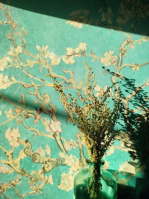 A green vase containing dried flowers casts intricate shadows against a textured, botanical-themed wall. The wallpaper features a vibrant teal background with a pattern of branches and blossoms, creating a harmonious blend of natural and artistic elements. Sunlight enhances the contrast and adds depth to the scene.