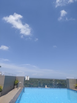 A rooftop swimming pool with clear blue water is surrounded by a concrete deck with metal railings. There are a few potted plants placed along the edge. The background features a panoramic view of lush green treetops extending towards the horizon under a clear blue sky with a few scattered white clouds.