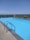 An outdoor swimming pool with clear blue water, surrounded by a concrete deck. A metal ladder is attached to the pool's edge for easy access. Green plants in decorative planters line the far side of the pool, with a safety glass barrier behind them. The background features a clear blue sky and a distant view of a lush green landscape.