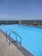 An outdoor swimming pool with clear blue water, surrounded by a concrete deck. A metal ladder is attached to the pool's edge for easy access. Green plants in decorative planters line the far side of the pool, with a safety glass barrier behind them. The background features a clear blue sky and a distant view of a lush green landscape.