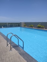 An outdoor swimming pool with clear blue water, surrounded by a concrete deck. A metal ladder is attached to the pool's edge for easy access. Green plants in decorative planters line the far side of the pool, with a safety glass barrier behind them. The background features a clear blue sky and a distant view of a lush green landscape.