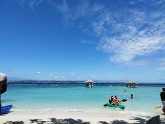 A serene beach scene with clear blue skies and turquoise water. Several people are enjoying the water and kayaking. Two thatched-roof structures are visible in the distance over the ocean. A kayak with two individuals is in the foreground, and more people are scattered across the beach and water.