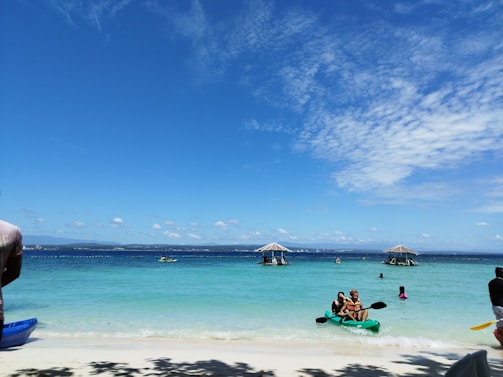 A serene beach scene with clear blue skies and turquoise water. Several people are enjoying the water and kayaking. Two thatched-roof structures are visible in the distance over the ocean. A kayak with two individuals is in the foreground, and more people are scattered across the beach and water.