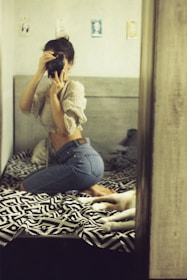 A person is kneeling on a bed taking a photo using a camera. The bed has a black and white patterned blanket, and there is a dog lying next to them. The room has a soft, warm lighting with light-colored walls and some artwork hanging on them.