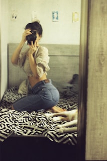 A person is kneeling on a bed taking a photo using a camera. The bed has a black and white patterned blanket, and there is a dog lying next to them. The room has a soft, warm lighting with light-colored walls and some artwork hanging on them.