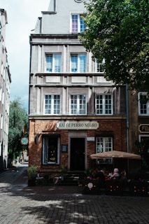 A quaint cafe located on a cobblestone street, housed in a multi-story building with large windows. The cafe has outdoor seating, shaded by a large umbrella, and is surrounded by potted plants and flowers. The facade of the building is a mix of brick and plaster, with a sign reading 'Kaffe Perro Negro'.