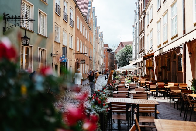 A charming European street lined with colorful buildings on each side, featuring traditional architectural designs. The street is paved with cobblestones and has numerous outdoor caf&eacute; tables, suggesting a bustling yet cozy atmosphere. People are seen walking, enjoying a leisurely day. Flower arrangements along the street add a touch of vibrancy.