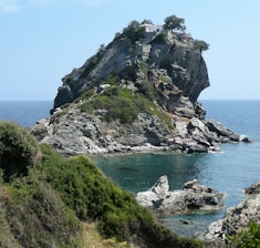 A rocky cliff rises dramatically from the turquoise sea, topped by a small white chapel with a Greek flag fluttering beside it. Green vegetation covers parts of the rock, contrasting with the gray tones of the stone. Clear blue sky and calm sea create a serene and picturesque seascape.