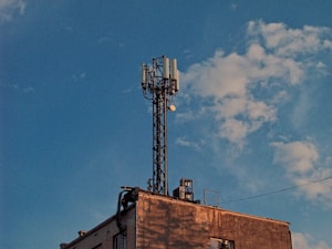 A telecommunications tower is mounted on the rooftop of a building, surrounded by a clear blue sky with scattered clouds. The structure is composed of various antennas and equipment.