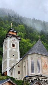 A picturesque church with a tall steeple situated against a backdrop of lush, green forested hills shrouded in mist. The church features traditional architecture with a prominent fresco on its side and a cross atop the steeple.