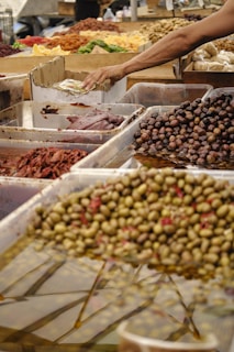 A variety of olives and pickled goods displayed in shallow water-filled trays at a market stall. A person is reaching to select an item, suggesting an interactive shopping experience. There are numerous containers filled with different types of olives, vegetables, and other preserved foods on the table.