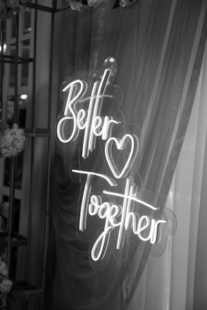A luminous neon sign displaying the words 'Better Together' with a heart symbol in between. The sign is surrounded by draped fabric and flowers, creating a romantic and intimate ambiance.