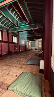 A traditional wooden interior with colorful ceiling beams, featuring intricate patterns and vibrant colors. The room contains several cushions on a polished wooden floor, suggesting a space for meditation or relaxation. Soft lighting filters through paper-covered windows, creating a serene and peaceful atmosphere.