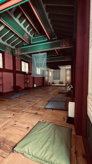 A traditional wooden intehttps://aryaflowyoga.com/wp-content/uploads/2024/06/woman-traveling-in-france-2023-11-27-05-16-47-utc_Easy-Resize.com_.jpgr with colorful ceiling beams, featuring intricate patterns and vibrant colors. The room contains several cushions on a polished wooden floor, suggesting a space for meditation or relaxation. Soft lighting filters through paper-covered windows, creating a serene and peaceful atmosphere.