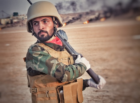 A man wearing military gear, including a camouflage jacket and protective helmet, is holding a rifle. The background is a blurred outdoor setting with a sandy terrain.