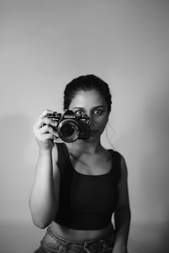 A monochrome image of a person holding a vintage Yashica camera at eye level, dressed in a black sleeveless top with a minimalist backdrop.