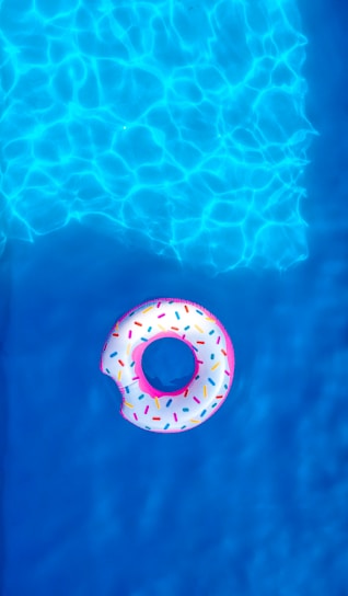A colorful inflatable pool float shaped like a donut with sprinkles is floating on the bright blue water. The sunlight creates dynamic patterns and reflections on the water surface.