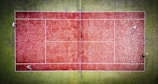 A bird's-eye view of a tennis court with a red surface surrounded by a green area. Four players are positioned on the court, each in a different corner, suggesting a doubles match in progress. The white lines on the court are clearly visible, marking the boundaries and net area. The contrast between the red court and green surroundings is striking, with the players adding dynamic elements to the composition.
