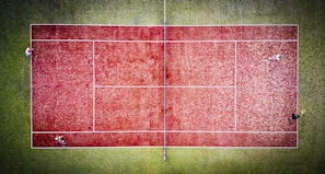 A bird's-eye view of a tennis court with a red surface surrounded by a green area. Four players are positioned on the court, each in a different corner, suggesting a doubles match in progress. The white lines on the court are clearly visible, marking the boundaries and net area. The contrast between the red court and green surroundings is striking, with the players adding dynamic elements to the composition.