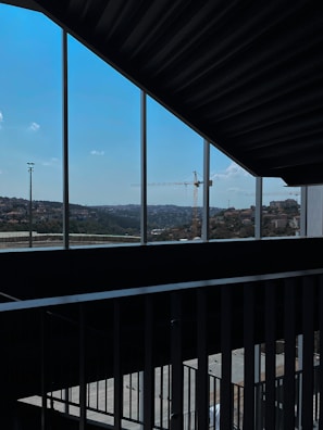 A view of a construction site, seen through a series of vertical window frames. A large crane is prominent against the skyline. The landscape outside includes rolling hills and a clear blue sky, while the interior features a metal railing and shadowed concrete surfaces.