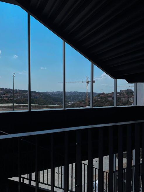 A view of a construction site, seen through a series of vertical window frames. A large crane is prominent against the skyline. The landscape outside includes rolling hills and a clear blue sky, while the interior features a metal railing and shadowed concrete surfaces.