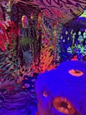 A vibrant and colorful room with abstract graffiti-like patterns covering the walls and ceiling. The space is illuminated with multicolored lights, creating an immersive and surreal atmosphere. There are hanging lamps, each encased in a textured translucent material, adding to the overall eclectic feel. In the foreground, a large blue and purple plush creature with an exaggerated face adds a whimsical element. In the background, a child can be seen interacting with the environment.