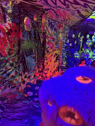 A vibrant and colorful room with abstract graffiti-like patterns covering the walls and ceiling. The space is illuminated with multicolored lights, creating an immersive and surreal atmosphere. There are hanging lamps, each encased in a textured translucent material, adding to the overall eclectic feel. In the foreground, a large blue and purple plush creature with an exaggerated face adds a whimsical element. In the background, a child can be seen interacting with the environment.