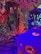 A vibrant and colorful room with abstract graffiti-like patterns covering the walls and ceiling. The space is illuminated with multicolored lights, creating an immersive and surreal atmosphere. There are hanging lamps, each encased in a textured translucent material, adding to the overall eclectic feel. In the foreground, a large blue and purple plush creature with an exaggerated face adds a whimsical element. In the background, a child can be seen interacting with the environment.