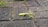 A bright green praying mantis is perched on a textured stone pathway with small patches of grass growing in the seams between the stones.